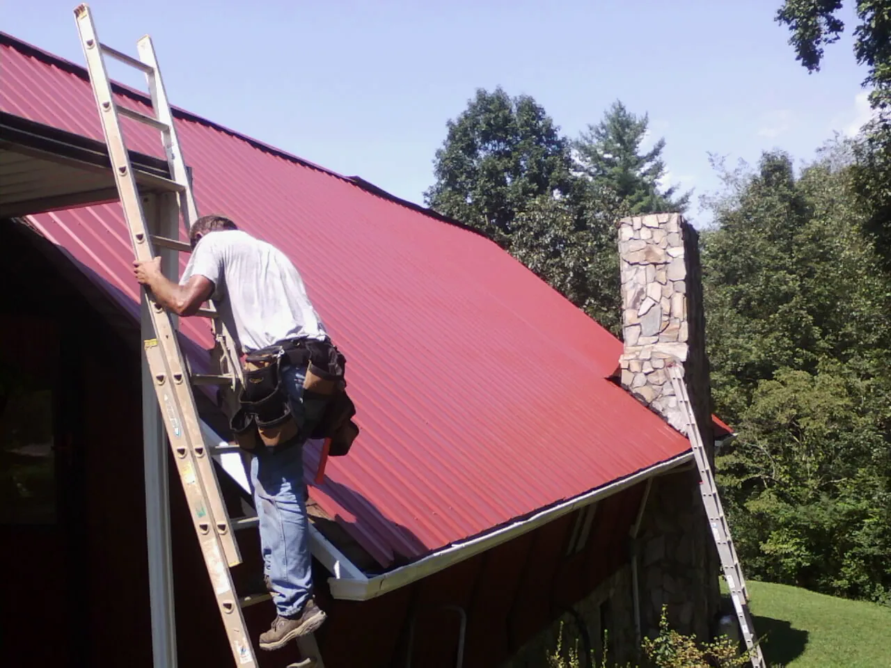 Roofer on a ladder beside a red metal roof and stone chimney — Soffit & Fascia Repair by Roofing Brothers in Alloy